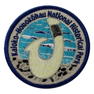 Round patch has name of Kaloko-Honokōhau National Historical Park embroidered around the edge of a scene depicting a white traditional bone fish hook against the blue and white ocean..