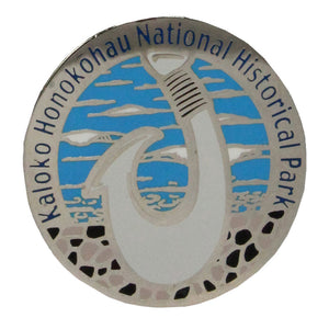 Pin shows a Hawaiian fish hook carved from bone or shell, a symbol of the advanced fishing techniques and achievement of early Hawaiian fishermen. Round enameled metal pin in blue, white and black is 1.25 x.75 inches in diameter and has the park name, Kaloko-Honokōhau National Historical Park, running around the top 3/4 edge of the pin.